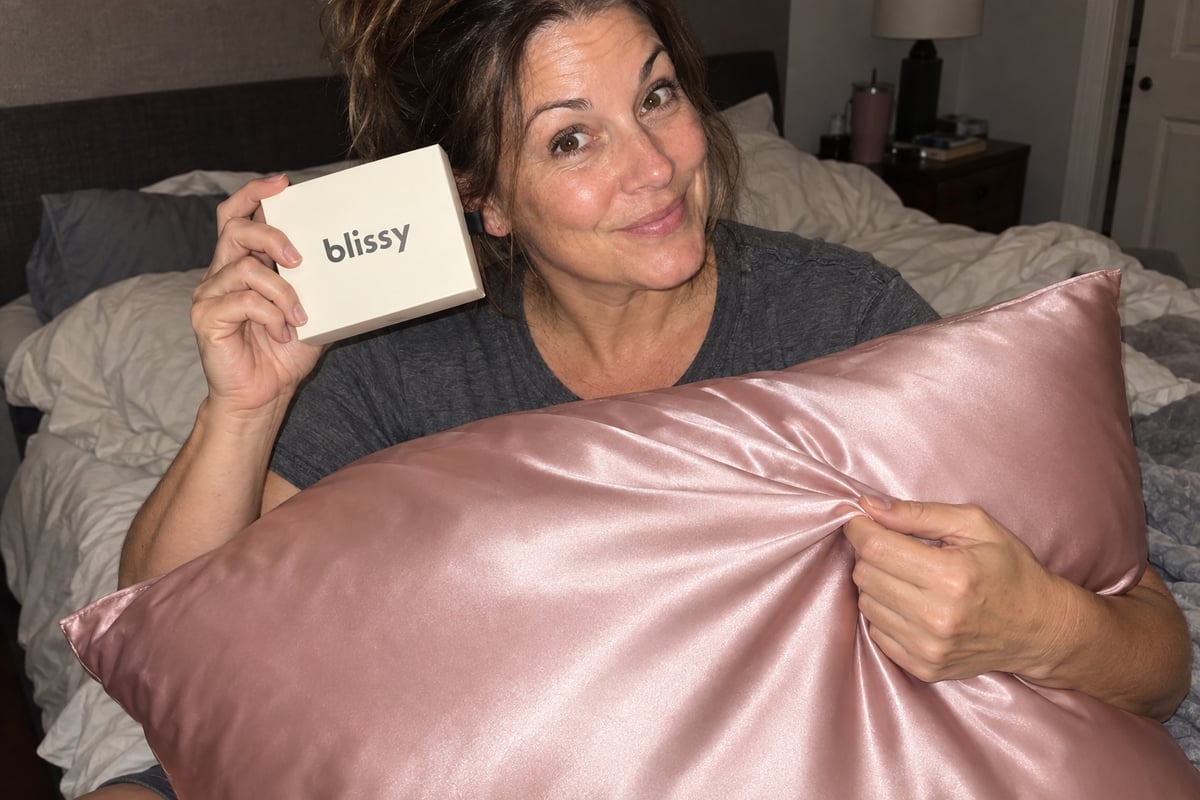 A woman holding a Blissy box and showing the fabric of a blush pillowcase on her pillow